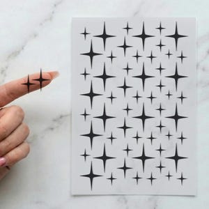May include: A rectangular sheet of white adhesive material featuring a repeating pattern of black four-pointed star shapes in various sizes. One individual black star sticker is shown detached from the sheet and placed on a fingertip.
