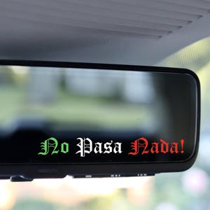 以下が含まれることがあります： 黒い車のバックミラーに、緑、白、赤のゴシック体で「No Pasa Nada！」と書かれたステッカー。ステッカーはミラーに貼られており、車の内装を映し出しています。