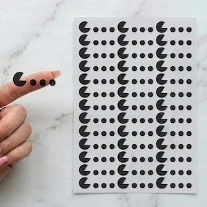 May include: Sheet of black Pac-Man stickers with a white background. A finger is shown with three Pac-Man stickers applied. The stickers are arranged in rows and columns, with each Pac-Man facing the same direction.