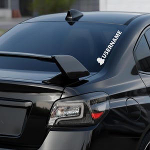 May include: A black car with a white decal on the rear window. The decal features a ghost-like logo and the word "USERNAME" in a vertical font. The car has a spoiler and a sleek, modern design.