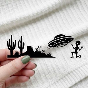 May include: A clear sticker featuring a black silhouette of a desert landscape with cacti, a UFO, and an alien. The alien is kneeling with its arms raised. The UFO is emitting a trail.