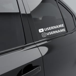 May include: White vinyl decal applied to a black car window. The decal features a YouTube play button icon and an Instagram logo, each followed by the word "USERNAME" in a sans-serif font. The car's door handle is visible.