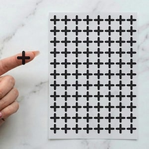 May include: A sheet of black plus sign stickers. A finger holds a single sticker, with a sheet of many stickers in the background. The stickers are on a white background.