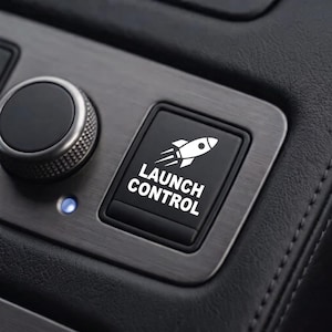 May include: A black square push button featuring a white graphic of a rocket ship and the text LAUNCH CONTROL in white capital letters. The button is set into a brushed metallic panel next to a circular dial and a small blue indicator light.