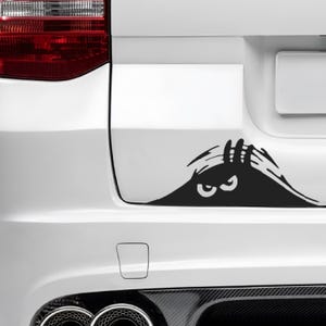 May include: Black vinyl car decal featuring a cartoon design of a peering monster with large eyes and clawed hands. The decal is applied to the white rear of a vehicle, adding a playful and unique touch.