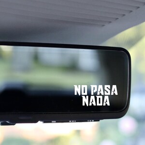 以下が含まれることがあります： 黒い車内バックミラーに、太字でスタイリッシュなフォントで「NO PASA NADA」と書かれた白いステッカーが貼られています。ステッカーは鏡面に直接貼られており、鏡には背景のぼやけた屋外の風景が映り込んでいます。