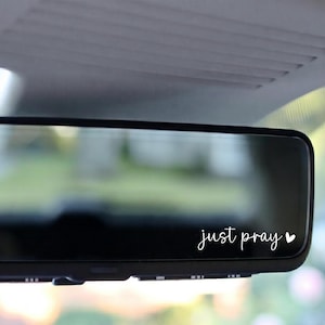May include: A black car rearview mirror with the words "just pray" in a white, cursive font. A small white heart is next to the word "pray". The mirror is set against a blurred background.