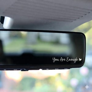 以下が含まれることがあります： 白い筆記体で「You Are Enough」と書かれた黒い車のバックミラー。テキストの横には小さな白いハートがあります。ミラーはぼやけた背景に対して設定されています。