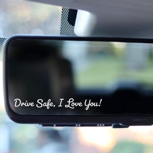 May include: A rectangular vehicle rearview mirror with a black frame. The surface of the mirror features white cursive text that reads, "Drive Safe, I Love You!" The mirror is mounted inside a vehicle, reflecting a blurred outdoor environment.