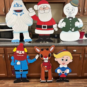 May include: A collection of six cardboard cutouts of popular Christmas characters. The characters include Santa Claus, a snowman, a Yeti, Rudolph the Red-Nosed Reindeer, an elf, and a lumberjack. The cutouts are standing on a countertop in a kitchen.