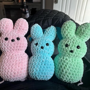 May include: Three handmade crochet bunny figures in pastel colors: pink, blue, and green. Each bunny has large ears, a rounded body, and black button eyes. The bunnies are made with a textured yarn, giving them a soft, plush appearance.