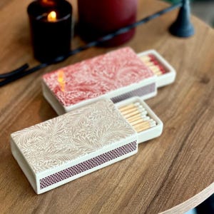 May include: Two rectangular matchboxes with sliding trays, one featuring a red and white botanical leaf pattern and the other a beige and white leaf pattern. Both boxes have a dark brown textured striking surface on the side and contain wooden matches.