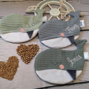 Könnte beinhalten: Drei walförmige Babykissen in Grün- und Grautönen, jedes mit einem Namen auf der Seite gestickt. Die Kissen haben ein strukturiertes Waffelmuster und einen weißen Bauch. Eine Herzform aus Samen ist ebenfalls sichtbar.