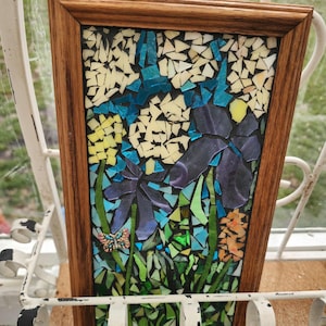 May include: A framed mosaic art piece featuring a floral design. The mosaic depicts purple irises, yellow and white flowers, and green foliage. A small butterfly is also included in the design. The frame is made of wood.