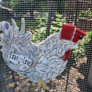 May include: A mosaic chicken sculpture with a red comb and beak, white body, and a tail made of newspaper clippings. The word "Inspire" is printed on the chicken's body. The sculpture is mounted on a wire mesh fence.