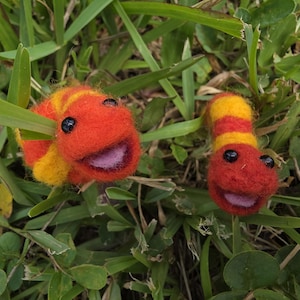 May include: Two needle-felted worm figures featuring horizontal stripes of vibrant orange and yellow. Each worm has a rounded head, small black bead eyes, and an open mouth revealing a light pink interior, positioned among green grass blades.
