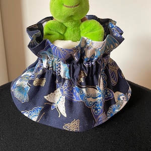 Könnte beinhalten: Ein dunkelblauer Stoffkragen mit einem gekräuselten Rand und einem Schmetterlingsdruck. Ein grünes Plüsch-Schildkrötenspielzeug sitzt oben auf dem Kragen.