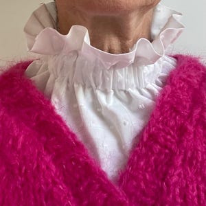 Könnte beinhalten: Ein weißer Rüschenkragen mit gerafftem Ausschnitt. Der Kragen wird über einem leuchtend pinkfarbenen, flauschigen Pullover getragen.