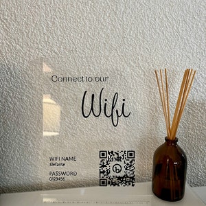 以下が含まれることがあります： Connect to our Wifi、WIFI NAME Elefante、PASSWORD 0123456という黒い文字とQRコードが印刷された透明なアクリル製サイン。その横には、木製のリードディフューザーのスティックが入った茶色のガラス瓶が白い表面の上に置かれています。