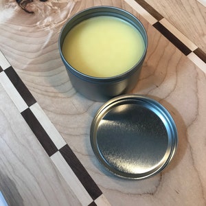 Puede incluir: Un recipiente de hojalata plateado con tapa, que contiene un bálsamo o cera amarilla. El recipiente está sobre una superficie de madera con un patrón de tablero de ajedrez.