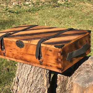 May include: A wooden box with a distressed finish and black leather straps and handles. The box has a metal latch and is sitting on a tree stump.