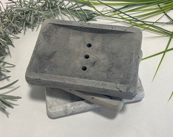 May include: A set of three gray concrete soap dishes. The top dish has three drainage holes. The bottom dish is white with gray marbling.