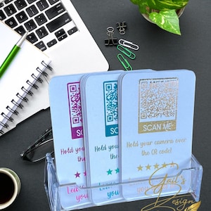 May include: A clear acrylic card holder with three cards featuring QR codes in purple, teal, and gold. Each card has text that reads "Hold your camera over the QR code!" and "Scan Me". The cards are on a black surface.
