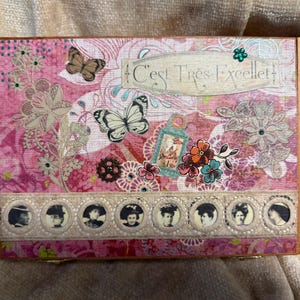 May include: A rectangular wooden box featuring a pink and white collage design with butterflies, floral patterns, and a gear. The text C'EST TRÈS EXCELLET is printed on a label. A horizontal strip displays eight circular portraits of individuals.