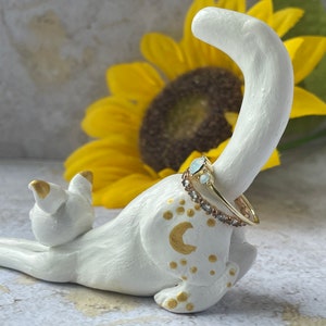 May include: White ceramic cat ring holder with gold accents and a ring with a light blue stone. The cat is sitting up with its tail curled around its body. The ring holder is in front of a yellow sunflower.