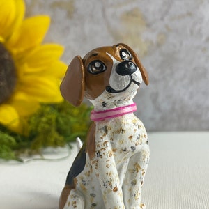 Puede incluir: Una figurita de cerámica pintada a mano de un perro beagle con pelaje marrón y blanco. El perro lleva un collar rosa y tiene una expresión juguetona en su rostro.