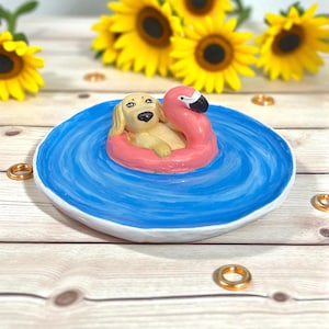 Puede incluir: Un plato de cerámica con interior azul y borde blanco presenta una figura de golden retriever flotando en un flotador de flamenco rosa. El plato está rodeado de girasoles amarillos y anillos dorados.