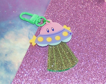May include: A green and purple UFO keychain with a cute face and yellow details. The keychain is on a sparkly pink background.