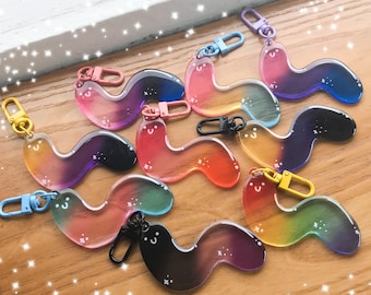 Puede incluir: Un conjunto de ocho llaveros coloridos con degradado arcoíris en forma de gusanos. Cada llavero tiene un anillo de metal plateado y una pequeña carita sonriente negra.