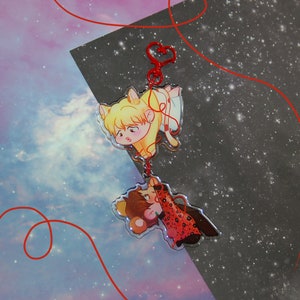 May include: A red string connects two cartoon characters. The first character is a blonde cat wearing a white shirt with a red bow. The second character is a brown cat wearing a red and black dress. Both characters are on a clear background with a galaxy pattern.