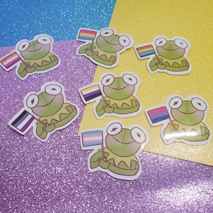 Pode incluir: Conjunto de 7 adesivos de sapo com diferentes bandeiras do orgulho. Cada adesivo apresenta um sapo verde segurando uma bandeira. As bandeiras incluem as bandeiras transgênero, bissexual, lésbica, pansexual, não binário, assexual e genderfluid.