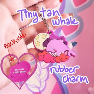 May include: A pink rubber charm keychain featuring a whale sleeping on a cloud with a moon. The back of the charm says "May you be a little happier".