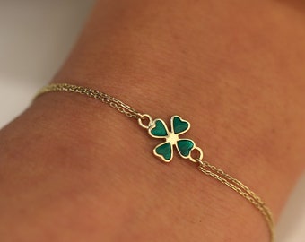 May include: A gold-colored bracelet featuring a four-leaf clover charm with green enamel. The bracelet has a delicate chain design and is worn on a wrist. The clover is a symbol of good luck.