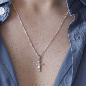 May include: A silver cross pendant necklace hangs against a blue button-down shirt. The cross is detailed with a small, decorative design. The necklace chain is delicate and silver. The shirt has white buttons.