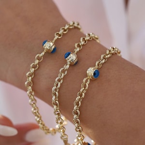 May include: Three gold chain bracelets with blue evil eye charms. The bracelets are layered on a person's wrist.