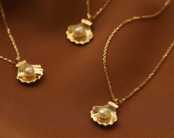Può includere: Tre collane d'oro con catene delicate e un ciondolo a forma di conchiglia con una perla. Le collane sono esposte su uno sfondo di tessuto marrone.