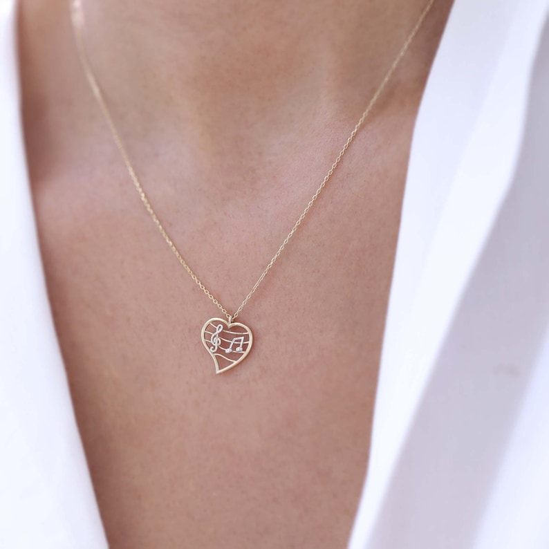 May include: A gold-colored necklace with a heart-shaped pendant. The pendant features a musical note design with a treble clef and musical notes. The necklace is worn against a white shirt.