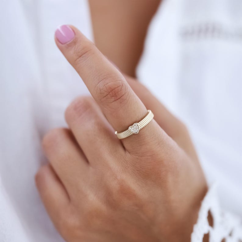 K&ouml;nnte beinhalten: Ein goldener Ring mit einem kleinen herzf&ouml;rmigen Diamanten in der Mitte. Der Ring befindet sich an einem Finger.