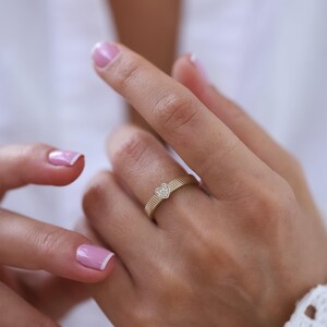 K&ouml;nnte beinhalten: Ein goldener Ring mit einem kleinen herzf&ouml;rmigen Diamanten in der Mitte. Der Ring hat eine strukturierte Band.