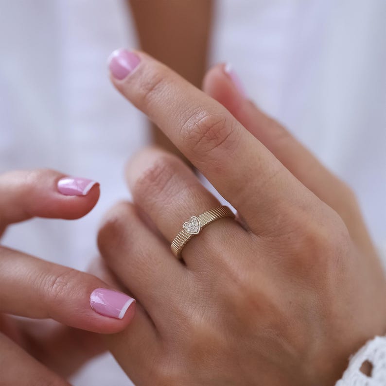 K&ouml;nnte beinhalten: Ein goldener Ring mit einem kleinen herzf&ouml;rmigen Diamanten in der Mitte. Der Ring hat eine strukturierte Band.