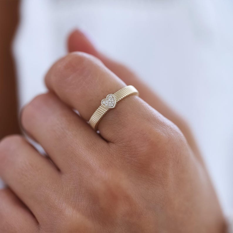 K&ouml;nnte beinhalten: Ein goldener Ring mit einem herzf&ouml;rmigen Diamanten in der Mitte. Die Band ist mit einem gerippten Design strukturiert.