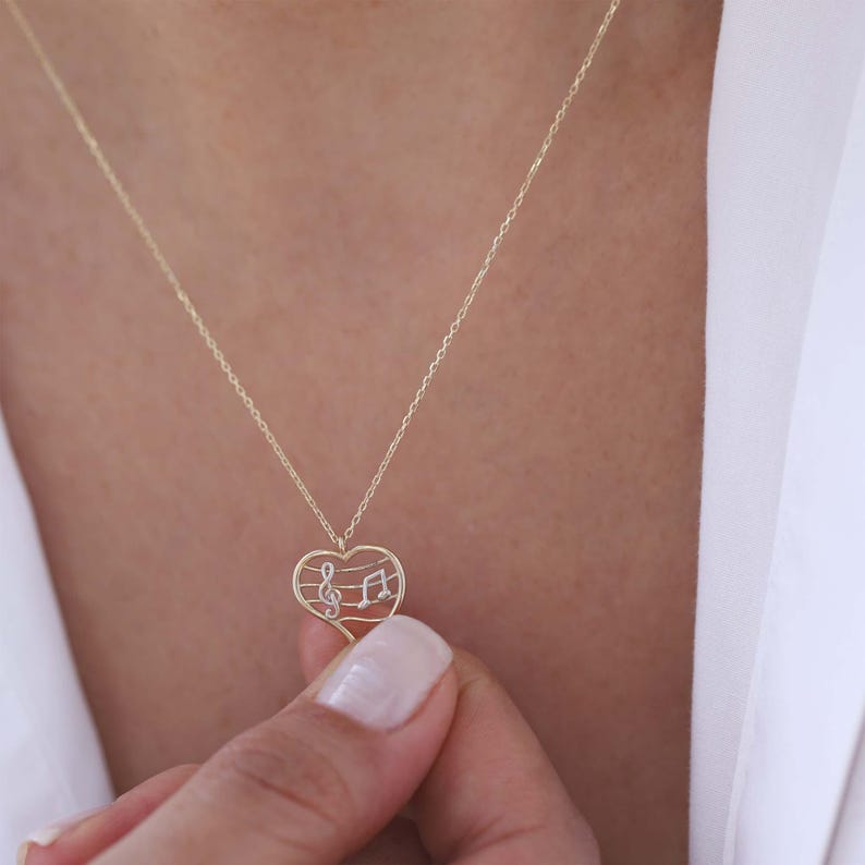 May include: A gold-colored necklace with a heart-shaped pendant. The pendant features a musical note design with a treble clef and a quarter note. The necklace is worn around the neck.
