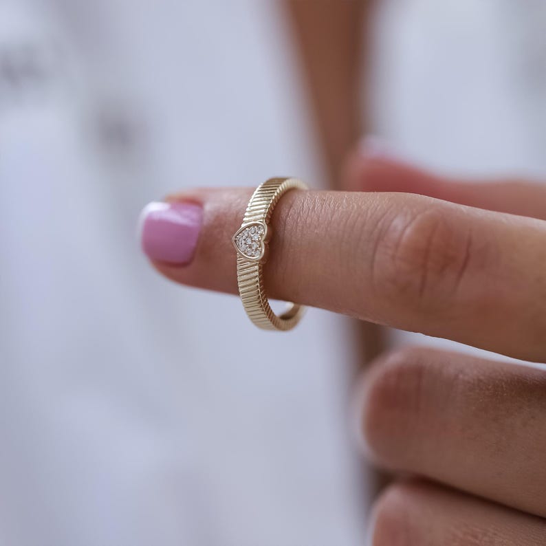 K&ouml;nnte beinhalten: Ein goldener Ring mit einem herzf&ouml;rmigen Diamanten in der Mitte. Der Ring hat eine strukturierte Band.