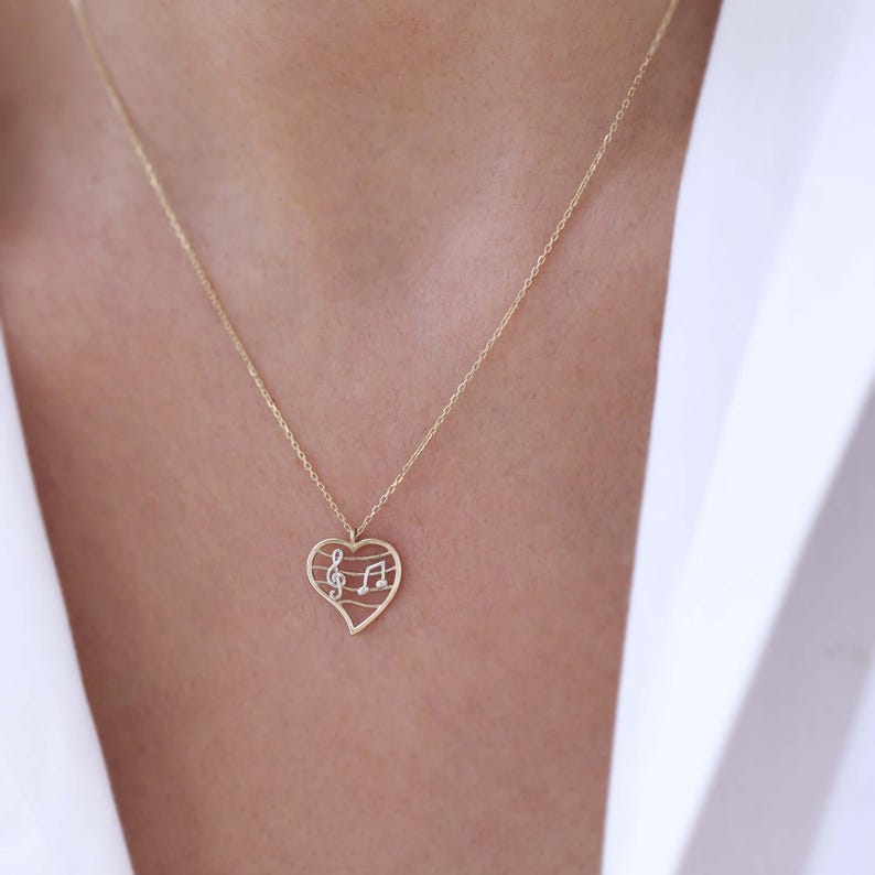 May include: A gold-colored necklace featuring a heart-shaped pendant. The pendant has a cutout design with musical notes and a treble clef. The necklace is worn around the neck, and the chain is delicate.