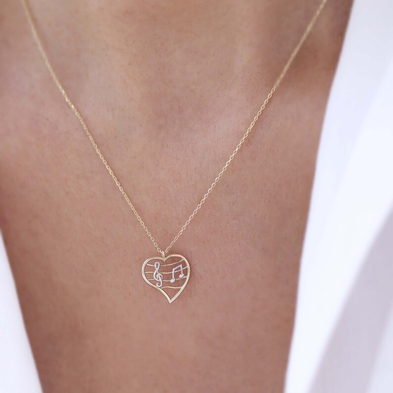 May include: A gold-colored necklace featuring a heart-shaped pendant. The pendant has a musical note design with a treble clef and musical notes. The necklace is delicate and appears to be made of a fine metal chain.