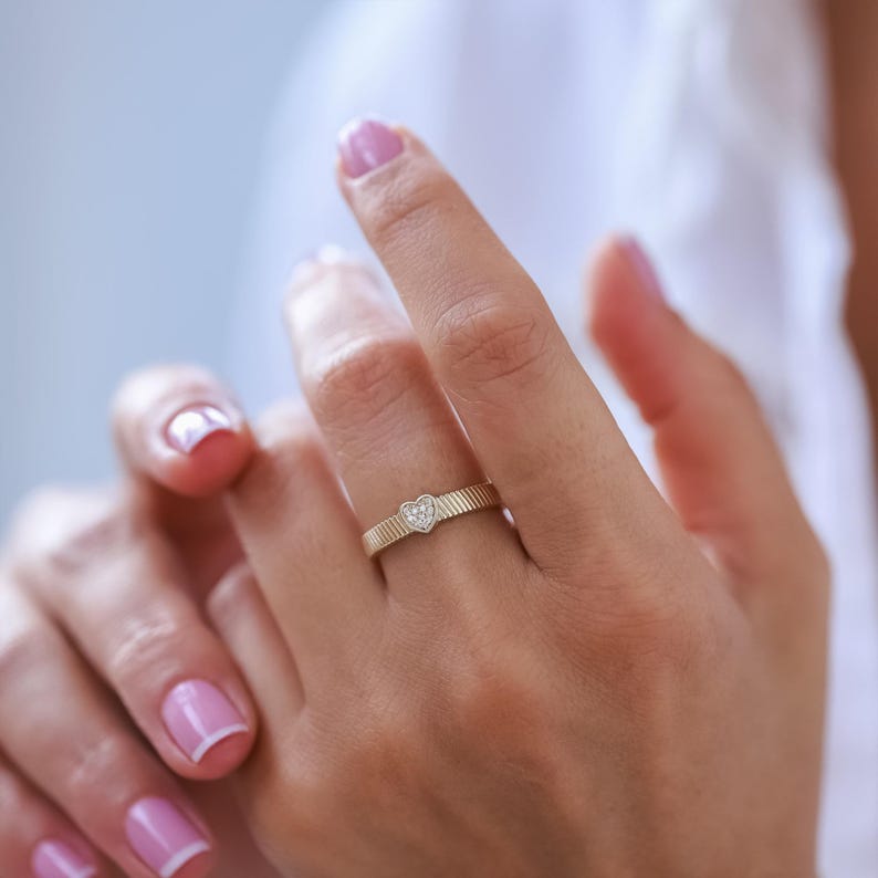 K&ouml;nnte beinhalten: Ein Goldring mit einem kleinen herzf&ouml;rmigen Diamanten in der Mitte. Der Ring befindet sich an einem Finger.